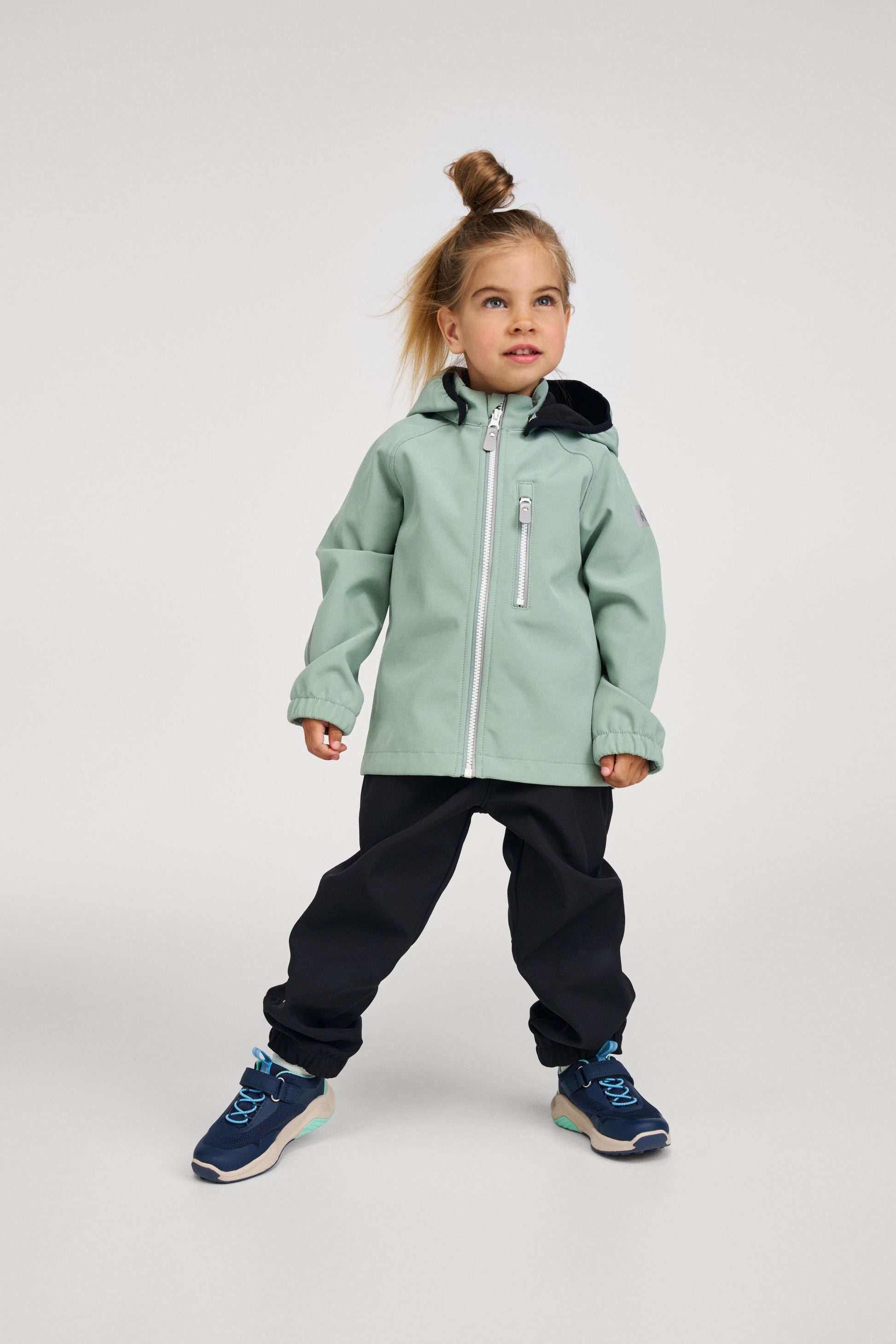 Lifestyle shot of a young girl wearing the stone green Reima Vantti kids' waterproof softshell jacket paired with black outdoor pants and blue sneakers, demonstrating its comfortable, active fit and stylish design for everyday outdoor play.