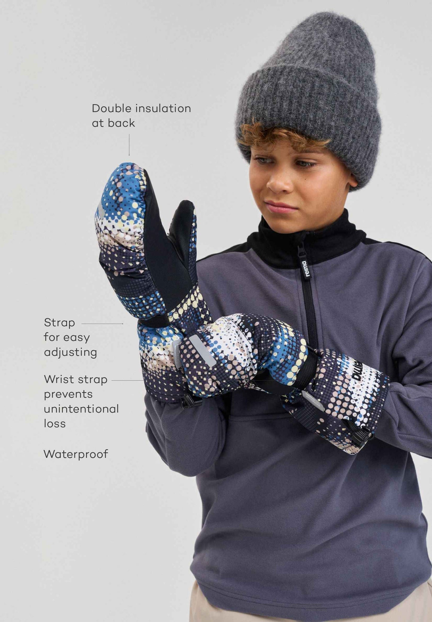 Front lifestyle view of a young skier wearing the soft black patterned Reima Vanukko children's snow mittens. The image demonstrates how the adjustable wrist straps and extended cuffs provide a secure, weatherproof seal for active winter sports.