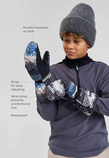 Front lifestyle view of a young skier wearing the soft black patterned Reima Vanukko children's snow mittens. The image demonstrates how the adjustable wrist straps and extended cuffs provide a secure, weatherproof seal for active winter sports.