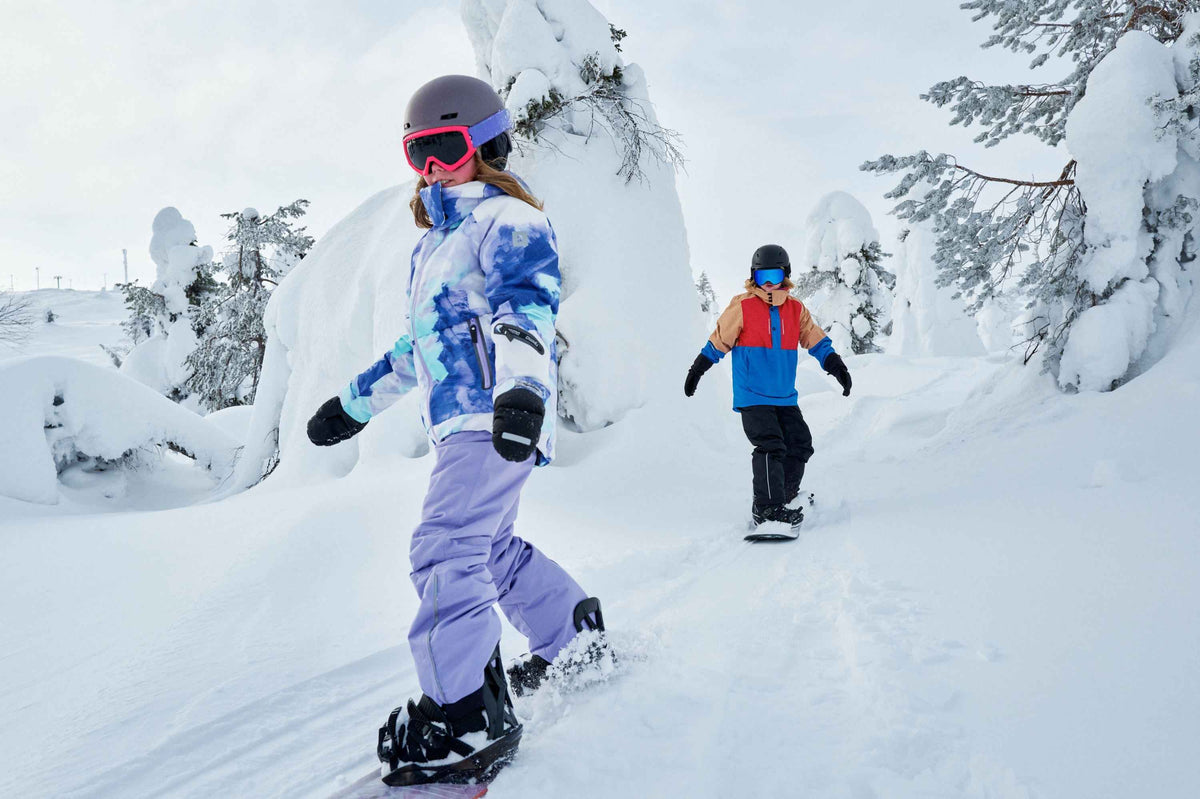Lifestyle action shot of a young girl snowboarding down a snowy mountain trail while wearing the waterproof black Reima Vanukko kids' winter mittens. She is geared up for alpine sports in a blue patterned winter jacket, purple snow pants, a grey helmet, and bright pink ski goggles, demonstrating the secure fit of the mittens during active winter play.
