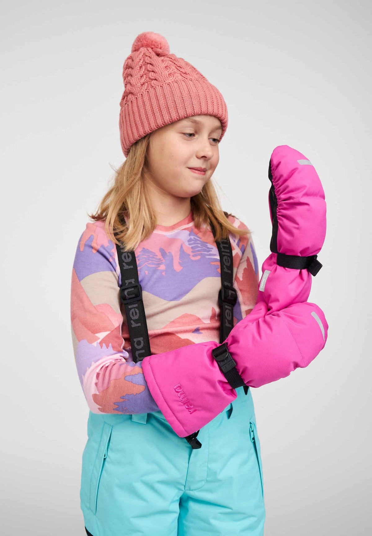 A young girl modeling the vibrant fuchsia Reima Vanukko kids' winter ski mittens, demonstrating the long protective cuffs and black palm grips. She is styled for winter sports in a pink knit beanie, patterned base layer, and light blue ski pants.