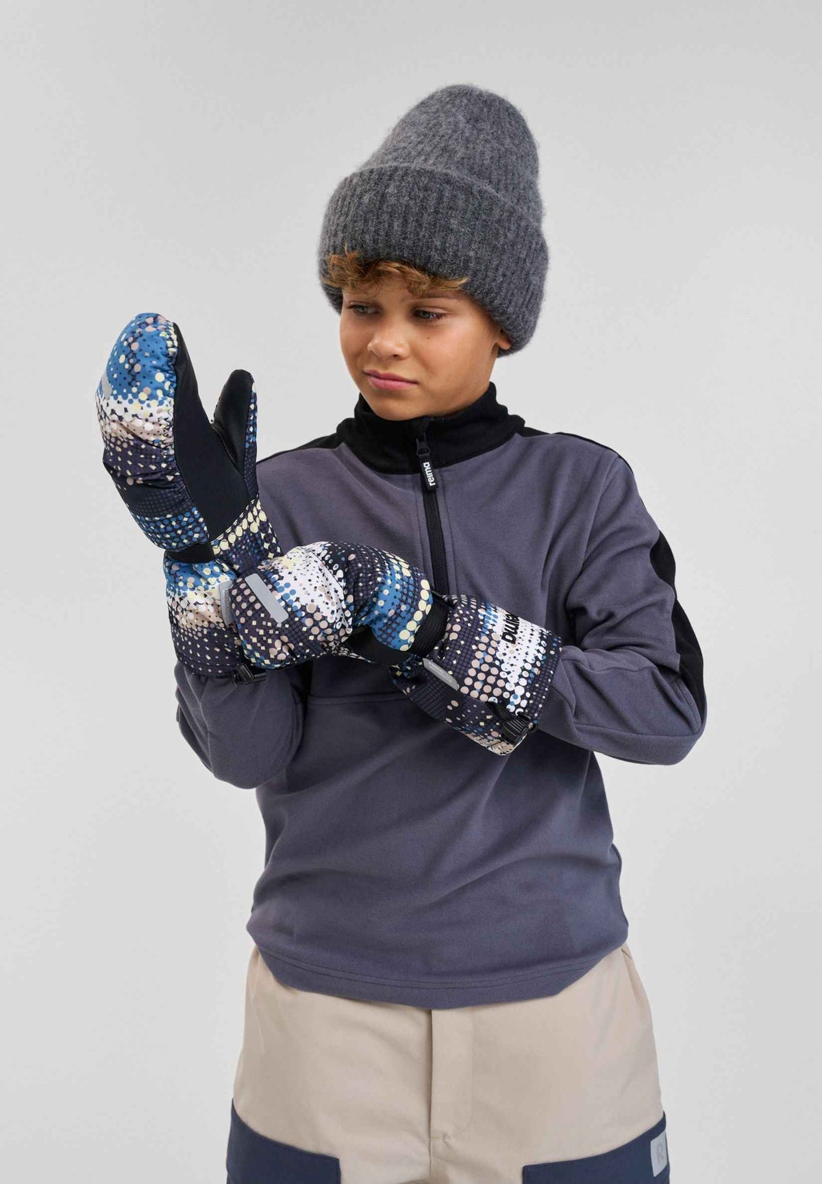 Lifestyle portrait of a young boy adjusting his soft black patterned Reima Vanukko kids' ski mittens. The image highlights the extra-long gauntlet cuffs designed to easily slide over winter jacket sleeves for maximum snow protection on the mountain.