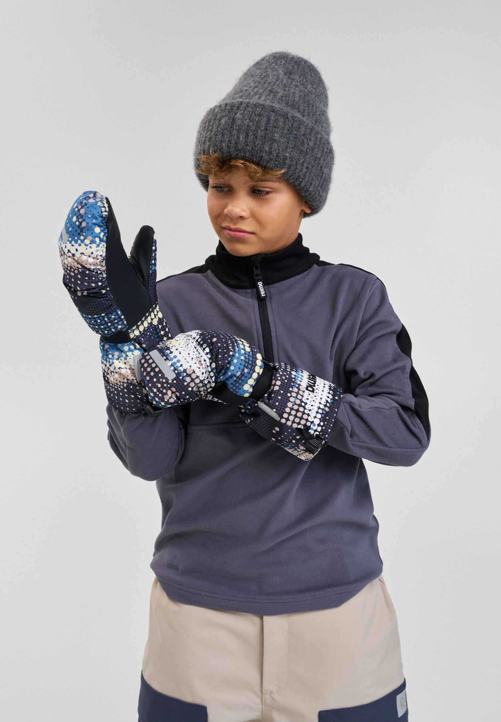 Lifestyle portrait of a young boy adjusting his soft black patterned Reima Vanukko kids' ski mittens. The image highlights the extra-long gauntlet cuffs designed to easily slide over winter jacket sleeves for maximum snow protection on the mountain.