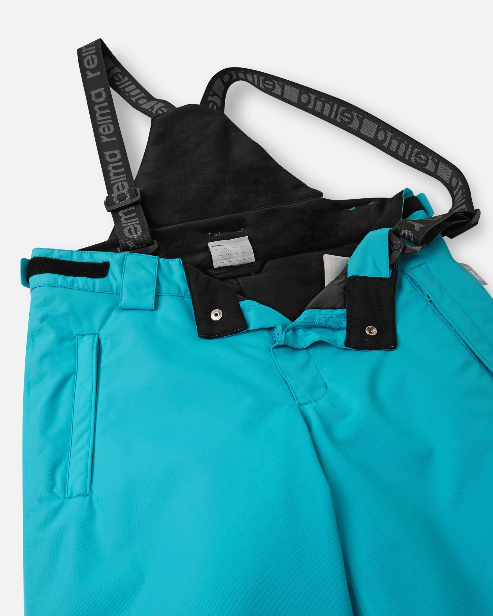 Close-up detail of the front waist on the aquatic Reima Wingon kids' ski pants, showing the secure snap buttons, smooth zipper fly, soft inner lining, and the front attachment points for the adjustable suspenders.