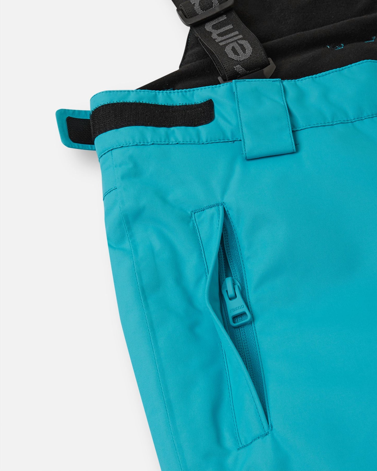 Close-up detail of the hip on the aquatic Reima Wingon kids' ski pants, highlighting a secure, low-profile zippered pocket and an adjustable hook-and-loop waist tab for a custom, draft-free fit.