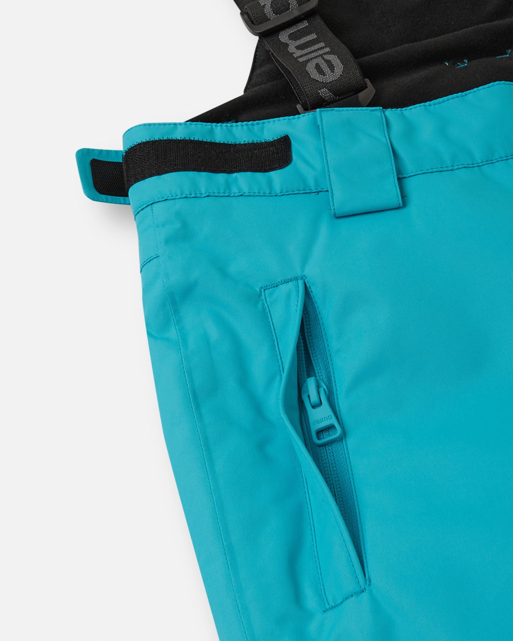 Close-up detail of the hip on the aquatic Reima Wingon kids' ski pants, highlighting a secure, low-profile zippered pocket and an adjustable hook-and-loop waist tab for a custom, draft-free fit.