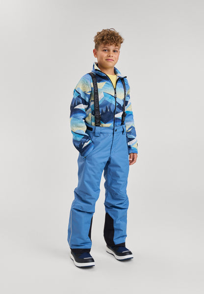 Lifestyle portrait of a young boy wearing the blue ocean Reima Wingon kids' winter ski pants paired with a patterned blue fleece jacket and snow boots. The image demonstrates the comfortable fit, mobility, and high-coverage design perfect for active snow play.