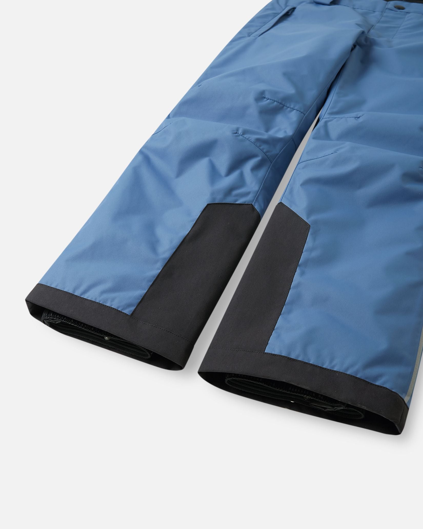 Detail view of the lower legs on the blue ocean Reima Wingon kids' ski pants, emphasizing the rugged black reinforced hems to protect against ski edges and the inner elastic snow locks designed to securely grip winter boots.