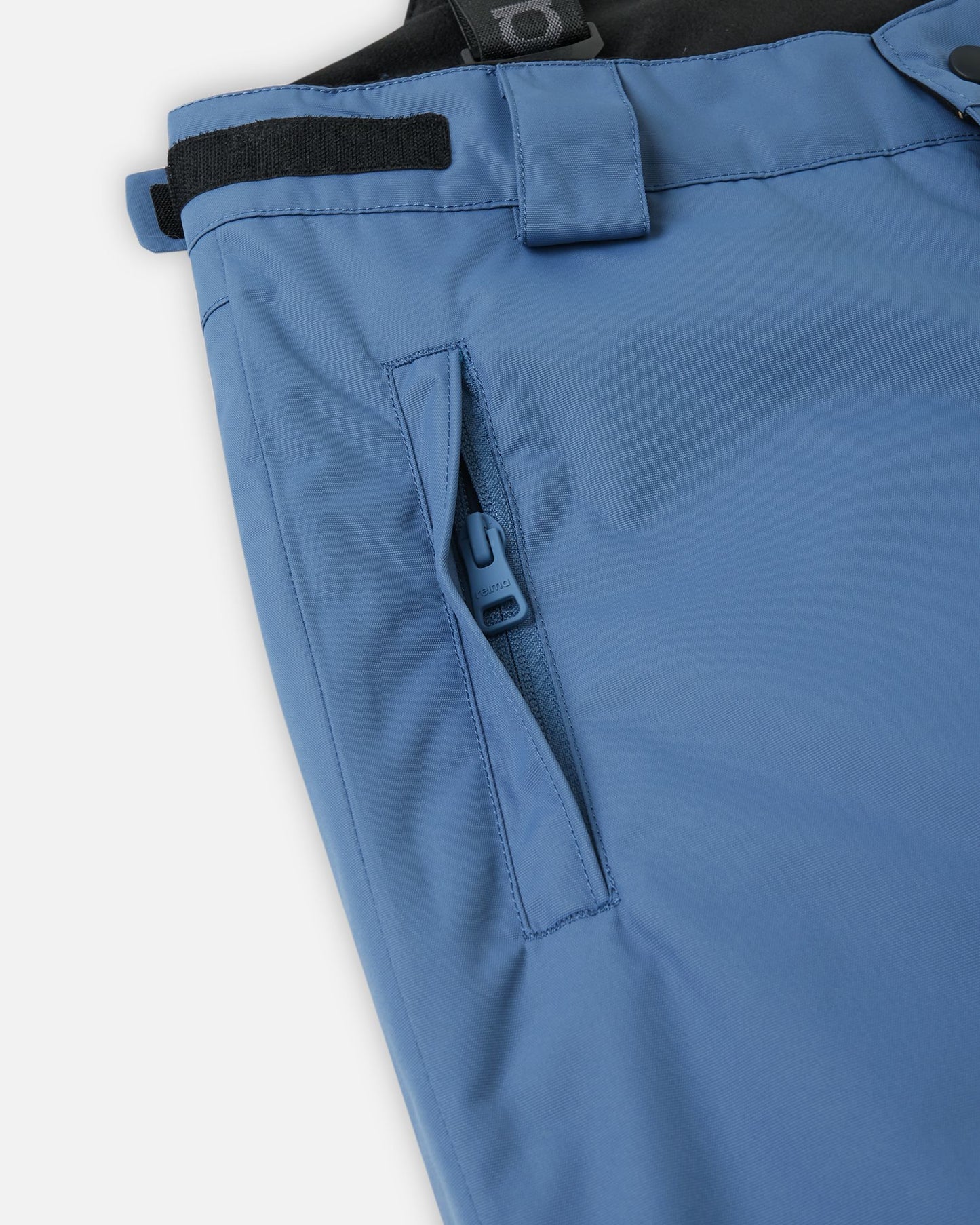 Close-up detail of the hip on the blue ocean Reima Wingon kids' ski pants, highlighting a secure, low-profile zippered pocket and an adjustable hook-and-loop waist tab for a custom, draft-free fit.