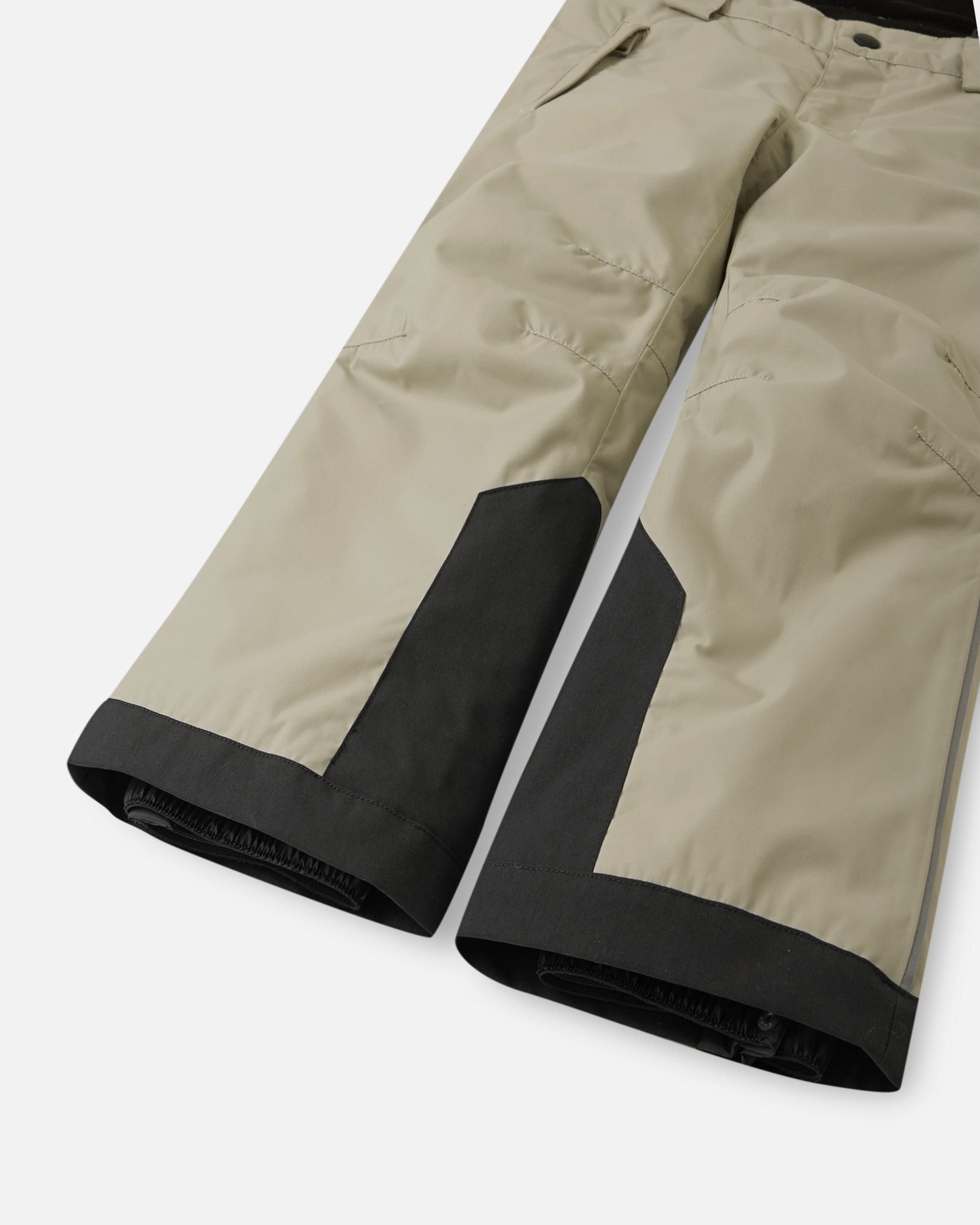 Detail view of the lower legs on the earthy beige Reima Wingon kids' ski pants, emphasizing the rugged black reinforced hems to protect against ski edges and the inner elastic snow locks designed to securely grip winter boots.