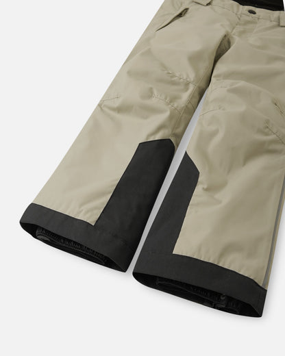 Detail view of the lower legs on the earthy beige Reima Wingon kids' ski pants, emphasizing the rugged black reinforced hems to protect against ski edges and the inner elastic snow locks designed to securely grip winter boots.