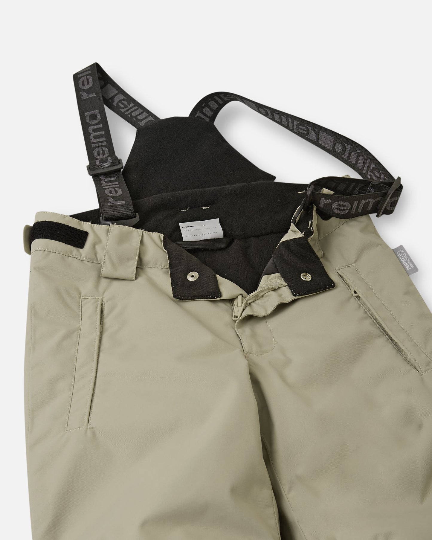 Close-up detail of the front waist on the earthy beige Reima Wingon kids' ski pants, showing the secure snap buttons, smooth zipper fly, soft inner lining, and the front attachment points for the adjustable suspenders.