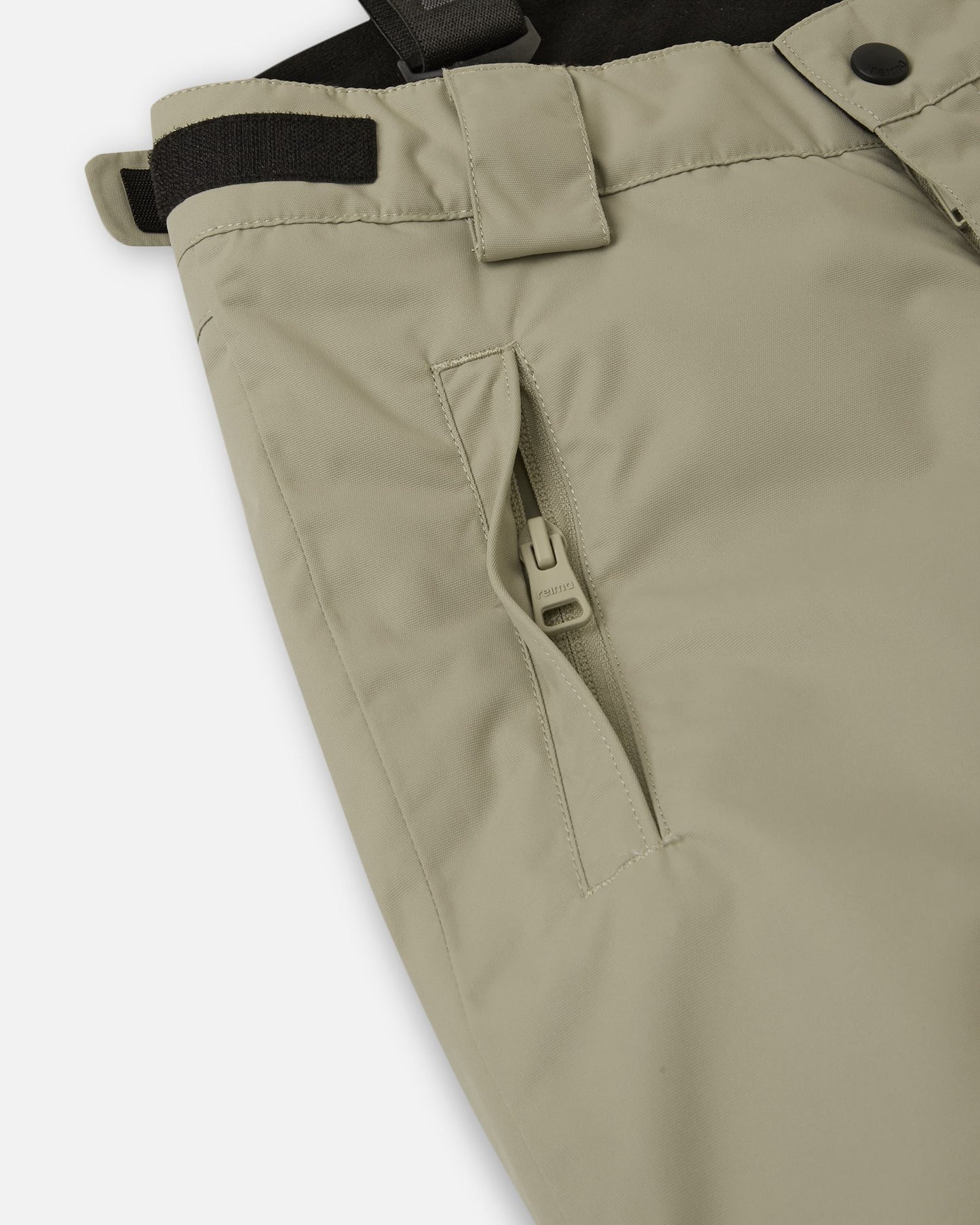 Close-up detail of the hip on the earthy beige Reima Wingon kids' ski pants, highlighting a secure, low-profile zippered pocket and an adjustable hook-and-loop waist tab for a custom, draft-free fit.