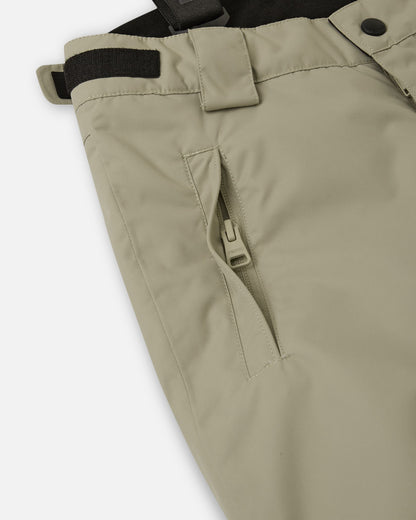 Close-up detail of the hip on the earthy beige Reima Wingon kids' ski pants, highlighting a secure, low-profile zippered pocket and an adjustable hook-and-loop waist tab for a custom, draft-free fit.