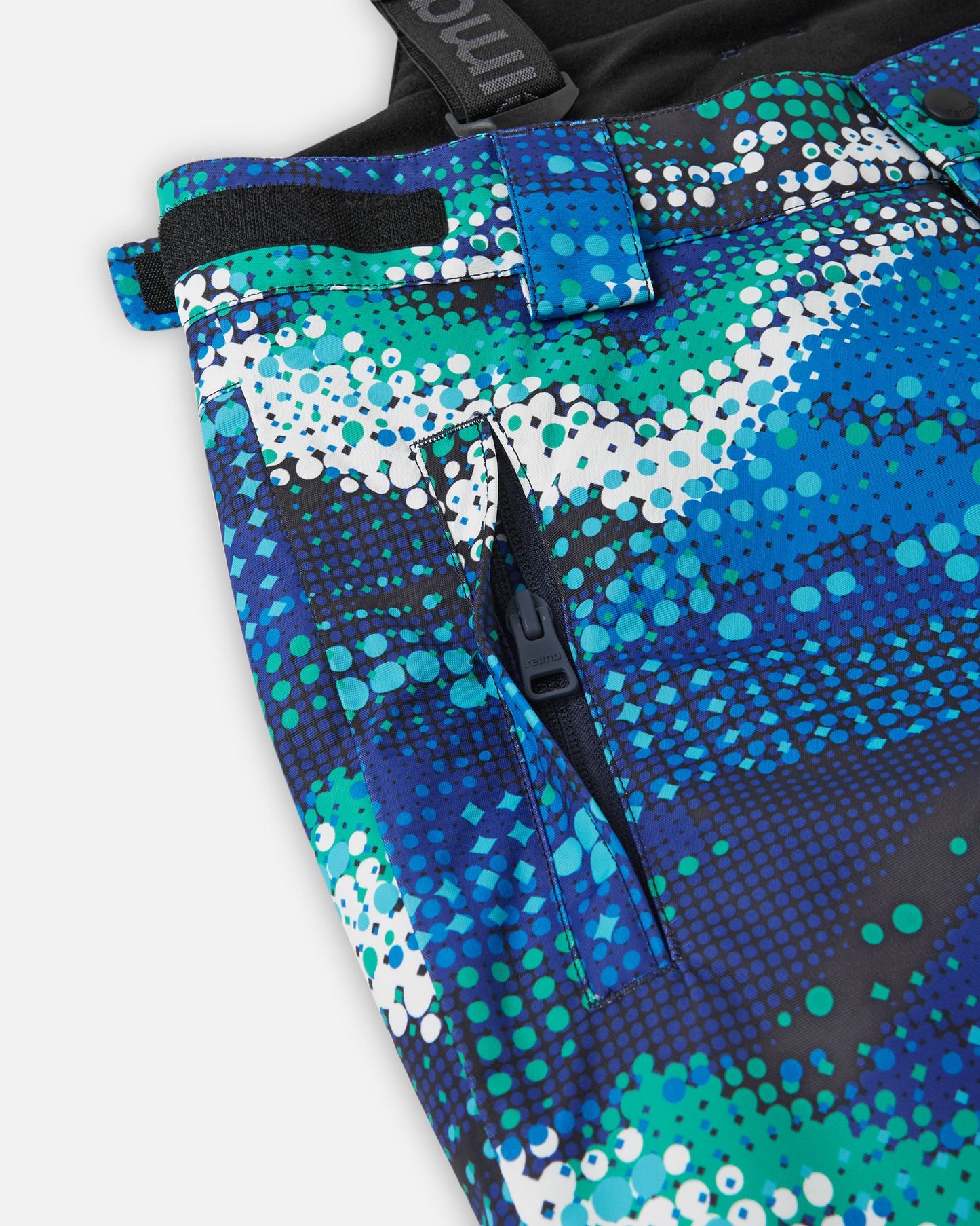 Close-up detail of the hip on the twilight blue Reima Wingon kids' ski pants, highlighting a secure, low-profile zippered pocket and an adjustable hook-and-loop waist tab for a custom, draft-free fit.